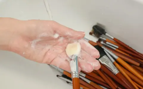 Mano con espuma jabonosa limpiando brochas de maquillaje en un lavabo.