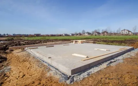 Solidny dom na płycie fundamentowej w budowie. W tle widać inne domy i zielone pola.