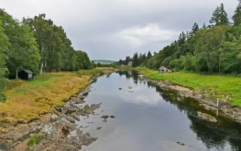 Ein ruhiger Fluss, der durch eine grüne Landschaft fließt. Kleine Häuser säumen das Ufer, vielleicht Orte, an denen man das schottische Uisge Beatha genießt.