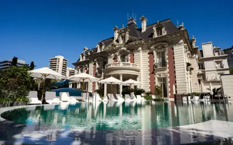 En lyxig pool framför en ståtlig byggnad som påminner om White Lotus Hotel, med solstolar och parasoller.