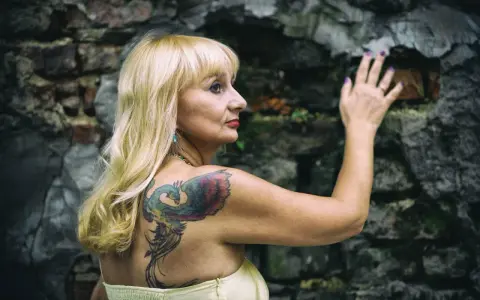 Blondynka z tatuażem feniksa na plecach opiera się o kamienną ścianę. Czyżby moda na tatuaże mijała, a ona wciąż pielęgnuje swoje?