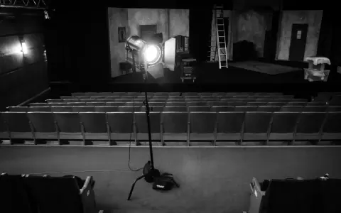 Teatr w czerni i bieli. Na scenie reflektor fresnel oświetla dekoracje, a przed nią puste rzędy siedzeń.