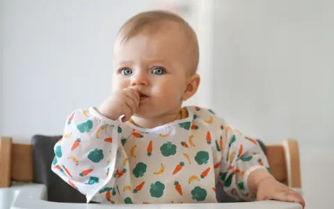 Niebieskooki maluch w śliniaczku z warzywami, kiedy dziecko rozpoznaje twarze, z paluszkiem w buzi.