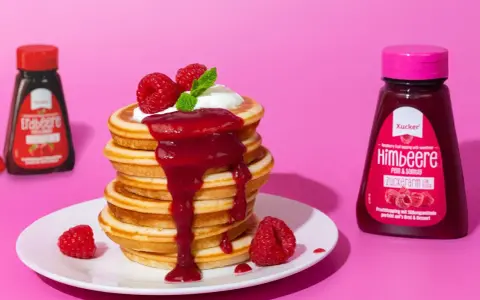 Ein Stapel zuckerfreie pancakes mit Himbeersauce und frischen Beeren. Daneben Flaschen mit Erdbeer- und Himbeer-Topping.