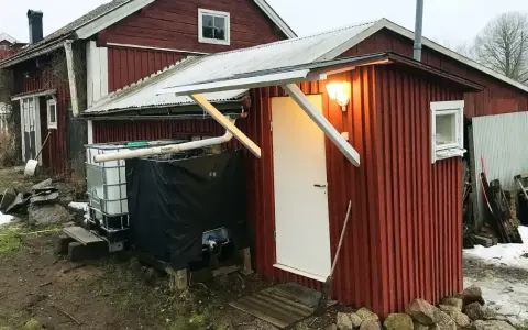 Rött utedass med vit dörr och fönster. Bredvid står en stor tank och en svart presenning. Här följer man utedass regler.