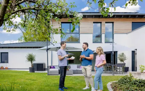 Tre personer diskuterar solcellslösningar på en grönskande trädgård framför ett modernt hus. PFA Solteknik hjälper dig att förverkliga dina drömmar.
