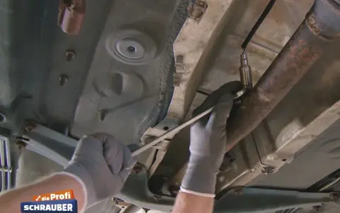 Ein Mechaniker löst die Lambdasonde am Auspuffrohr. Sie sitzt im vorderen Teil des Rohrs, nahe am Motor.