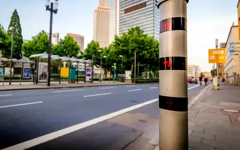 Blitzlicht warnt: Innerorts 17 kmh zu schnell. Ein Blitzer steht am Straßenrand, im Hintergrund Hochhäuser und Bushaltestellen.