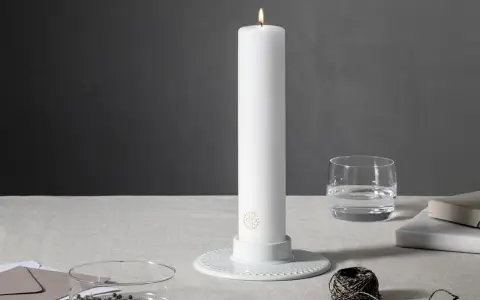 Ett vitt, högt stearinljus med guldig logotyp står i en vit ljusstake. Bredvid finns ett glas vatten, böcker och garn. Olika former av stearinljus kan skapa stämning.