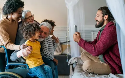 Familia multigeneracional reunida, una abuela en silla de ruedas, sonríen mientras un hombre toma una foto. Se hereda el Parkinson, pero el amor familiar perdura.