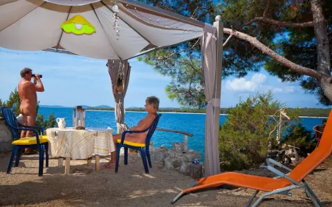 Para na plaży, relaksująca się w namiocie. Mężczyzna obserwuje morze przez lornetkę. To idealne miejsce na naturystyczny camping.