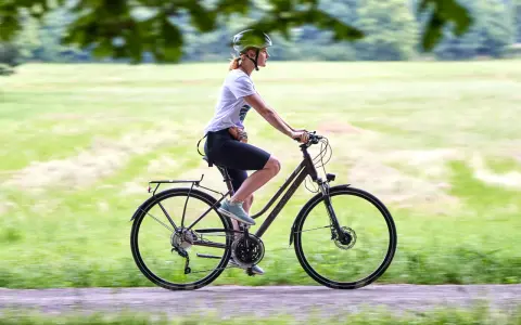 Kobieta w kasku jedzie na rowerze, który może być zarówno rowerem crossowym, jak i trekkingowym, po ścieżce w parku.