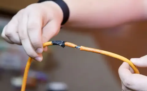 Dłonie trzymające pomarańczowy kabel z odsłoniętymi żyłkami, prowizorycznie połączone taśmą. Pokazuje, jak połączyć kable bez lutowania.