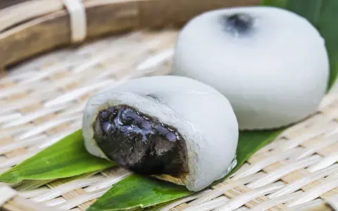 Dwa białe mochi, jedno przekrojone, ukazujące ciemne nadzienie. Podane na bambusowej tacce z liśćmi.
