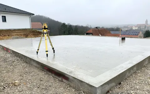 Der **Bodenplatten-Aufbau** ist fast fertig. Ein Nivelliergerät auf einem Stativ steht auf der frisch gegossenen Betonplatte. Im Hintergrund sind Häuser und eine Kirche zu sehen.