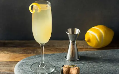 Elegancki drink z szampanem, ozdobiony skórką cytryny, obok kieliszka stoi miarka barmańska, cytryna i korek od szampana.