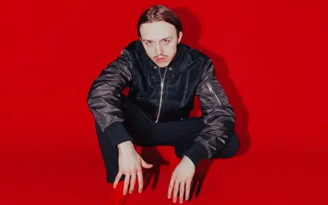 Tommy Cash w czarnej kurtce bomberce i czarnych spodniach kuca na czerwonym tle.