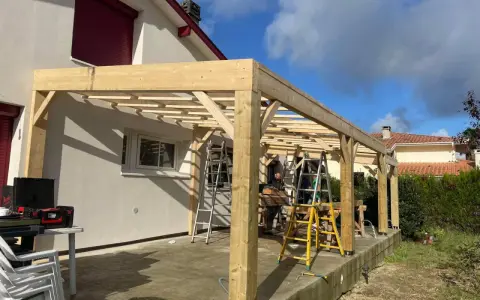Construction d'une charpente sans ferme en bois, avec des poteaux et des poutres, sur une terrasse. Des échelles sont présentes.