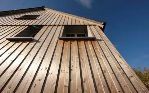 Vue en contre-plongée d'une maison avec un bardage bois clair, mettant en valeur la pose verticale des lames. Trois fenêtres sont visibles sous un ciel bleu éclatant.