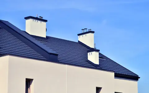 Nowoczesny dach bezokapowy z ciemnogranatową dachówką i białymi kominami na tle błękitnego nieba.