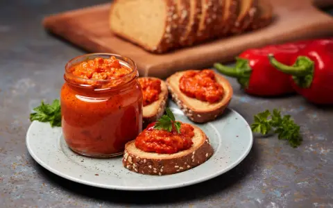 Słoik pełen ajvaru, pysznej pasty z papryki, podany z chlebem i świeżymi paprykami. Ajvar co to? To smak Bałkanów!