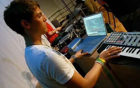 Junger Mann am Laptop und Keyboard, der Musik produziert. Ein Schlagzeuger im Hintergrund. Perfekt für ein Musikproduktion Studium.