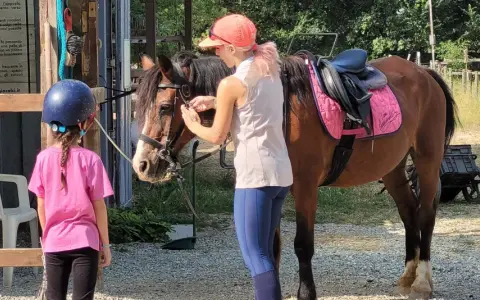 Bambina con casco blu e maglietta rosa osserva un cavallo marrone pronto per un giro a cavallo per bambini. Una ragazza con cappellino arancione prepara il cavallo.