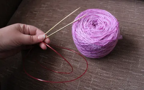 En hand håller i en rundsticka med ett nystan i lila toner bredvid. Dags att sticka resår på rundsticka!