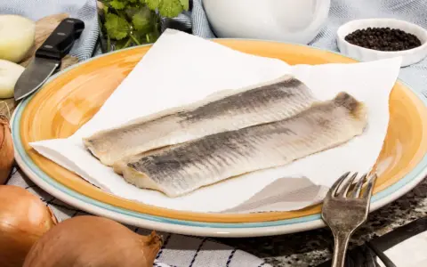 Zwei Matjesfilets auf Küchenpapier, bereit für eine gesunde Mahlzeit. Zwiebeln und Pfefferkörner deuten auf die Zubereitung hin.