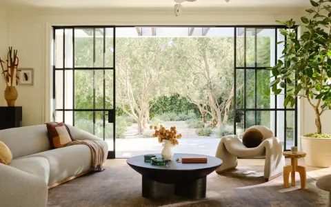 Salones modernos con sofá curvo, sillón acogedor y mesa de centro, conectados a un jardín soleado con olivos.