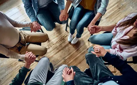 Grupa osób trzyma się za ręce w kręgu, wspierając się nawzajem. Widać tu pracę specjalisty od uzależnień.