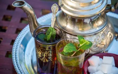 Zwei Gläser mit Minztee, garniert mit frischen Minzblättern, stehen neben einem traditionellen Teekessel und Zuckerwürfeln. Verschiedene Minze Arten verfeinern das Getränk.