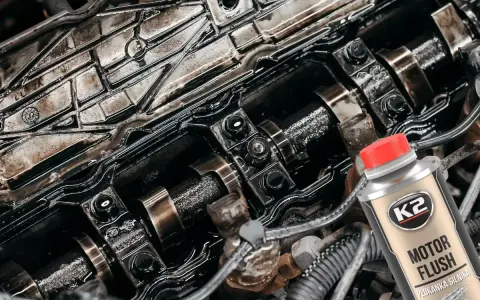 Silnik z widocznymi elementami rozrządu, pokryty czarnym osadem. Obok butelka płukanki K2 Motor Flush, która pomoże rozwiązać problem niskie ciśnienie oleju na rozgrzanym silniku.