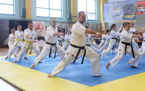 Grupa osób w kimonach ćwiczy karate na niebiesko-żółtej macie w sali gimnastycznej.