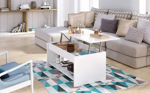 Mesa elevable pequeña blanca con compartimentos, junto a un sofá gris y una silla azul, sobre una alfombra geométrica.
