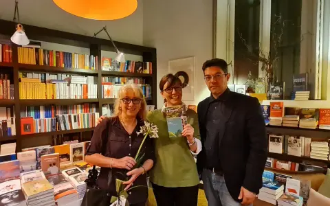 Tre persone sorridono in una libreria, una tiene in mano il libro "Cocco e Magella". Libri in ordine cronologico sugli scaffali.