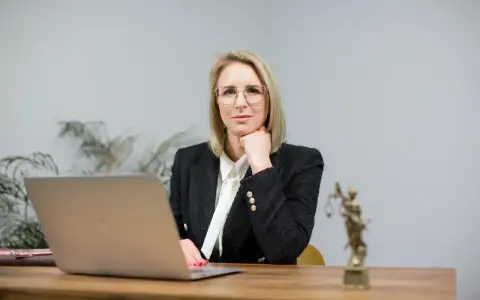Kobieta w okularach i żakiecie siedzi przy biurku z laptopem i statuetką Temidy.