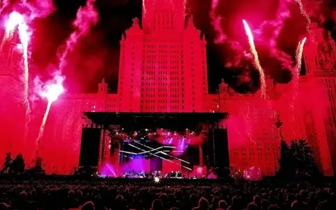 Ogromny budynek oświetlony na czerwono, fajerwerki i scena koncertowa. Tłum ludzi czeka na największy koncert na świecie.