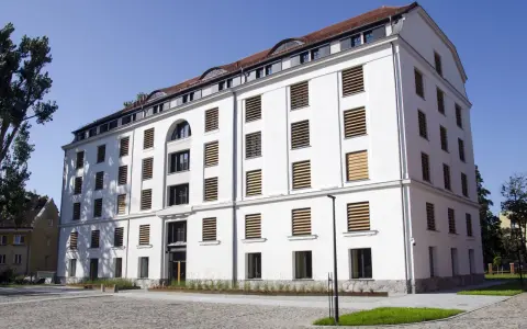 Nowoczesny, biały spichlerz w Słupsku z licznymi oknami o drewnianych żaluzjach, stojący na brukowanym placu.
