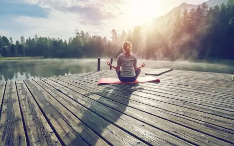 En kvinna med ryggen mot kameran sitter i meditationsställning på en brygga vid en sjö. Solen strålar genom dimman och skapar en magisk stämning. Hon verkar vara helt i nuet.