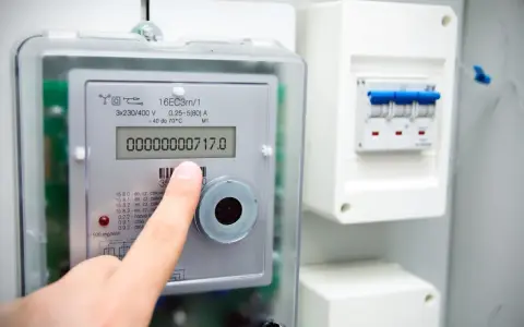 Palec wskazuje na cyfrowy licznik energii elektrycznej, pokazujący odczyt 00000000717.0. Widoczne są symbole na liczniku energii elektrycznej.