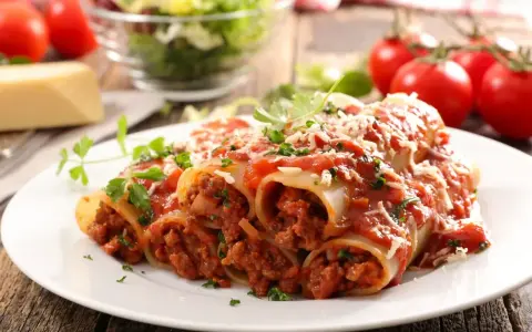 Pyszne cannelloni z mięsem mielonym, polane sosem pomidorowym i posypane parmezanem, z dodatkiem świeżej natki pietruszki.