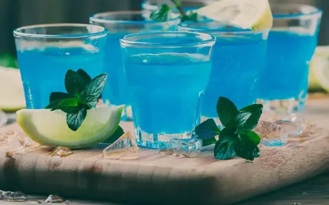 Kilka shotów z drinkiem blue curacao, ozdobionych limonką i miętą, na drewnianej desce z lodem.