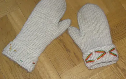 Två vita, stickade vantar med diskreta drops design-mönster i färggranna trådar på muddarna.