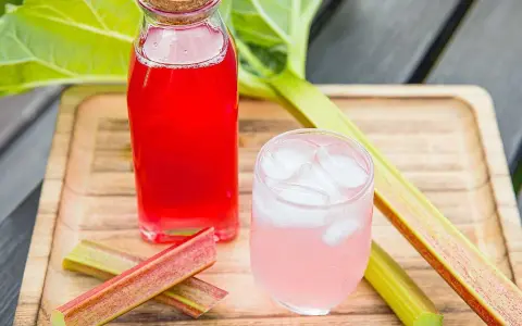 En flaska med röd rabarbersaft och ett glas med is och rosa rabarbersaft står på en träbricka. Rabarberstjälkar ligger bredvid.