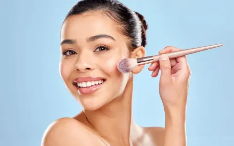 Une femme souriante utilise un pinceau pour appliquer du correcteur maquillage sur sa joue.