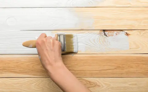 Ręka maluje białą farbą drewnianą boazerię, pokazując, jak odnowić starą boazerię.