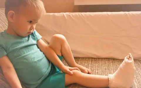 Chłopiec w turkusowej koszulce z nogą w gipsie siedzi na kanapie.