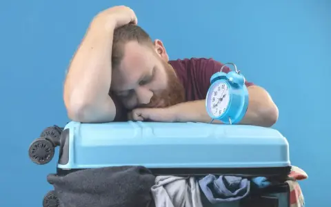 Zmęczony podróżnik śpi na walizce, budzik obok sygnalizuje jetlag.