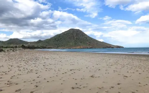 Weite Sandfläche am Playa de los Genoveses mit sanften Wellen und einem grünen Berg im Hintergrund unter einem bewölkten Himmel.